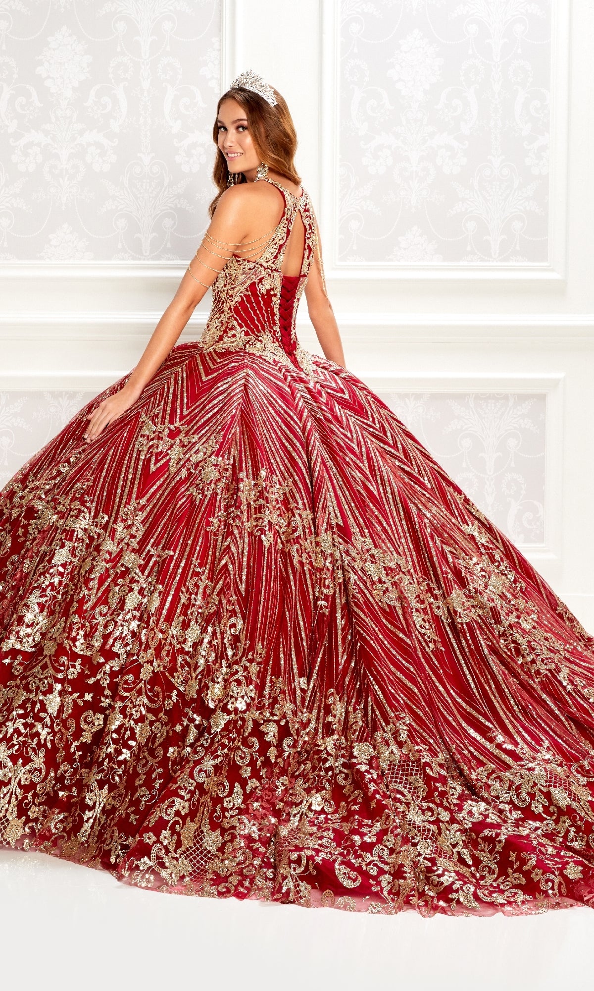 A woman models the Princesa PR22034 wine red quinceanera dress with gold embellishments and intricate patterns, posing in an ornate white room.